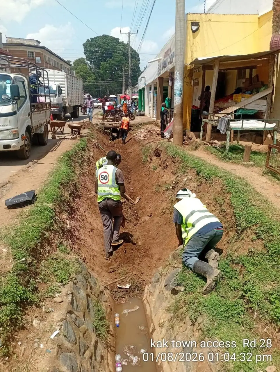 Two crew members excavating a roadside drain along a busy Kakamega trading street
