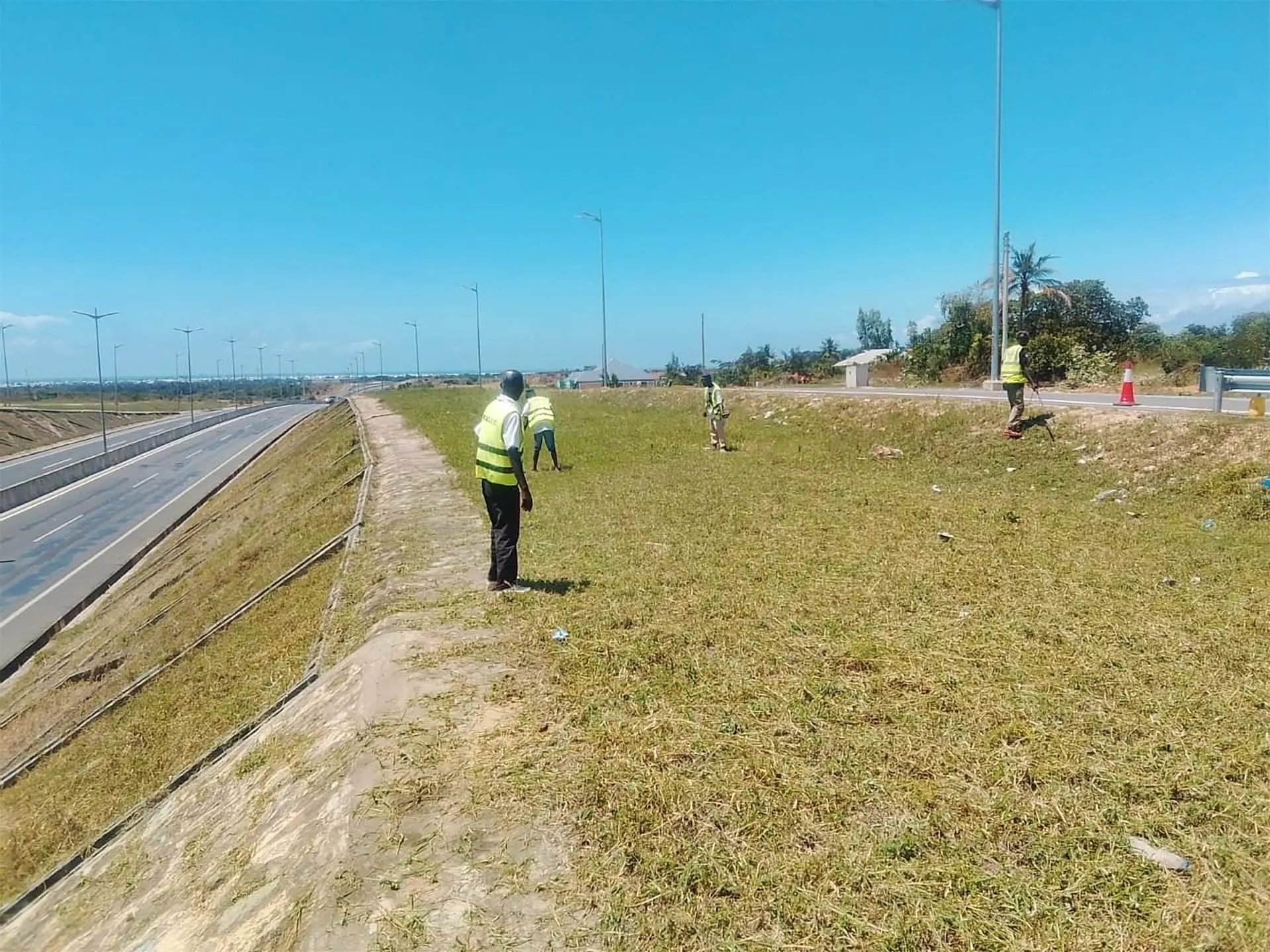 A newly maintained dual-carriageway section of the Mteza–Kibundani road, with a Kerada crew tending the embankment and the coastline beyond