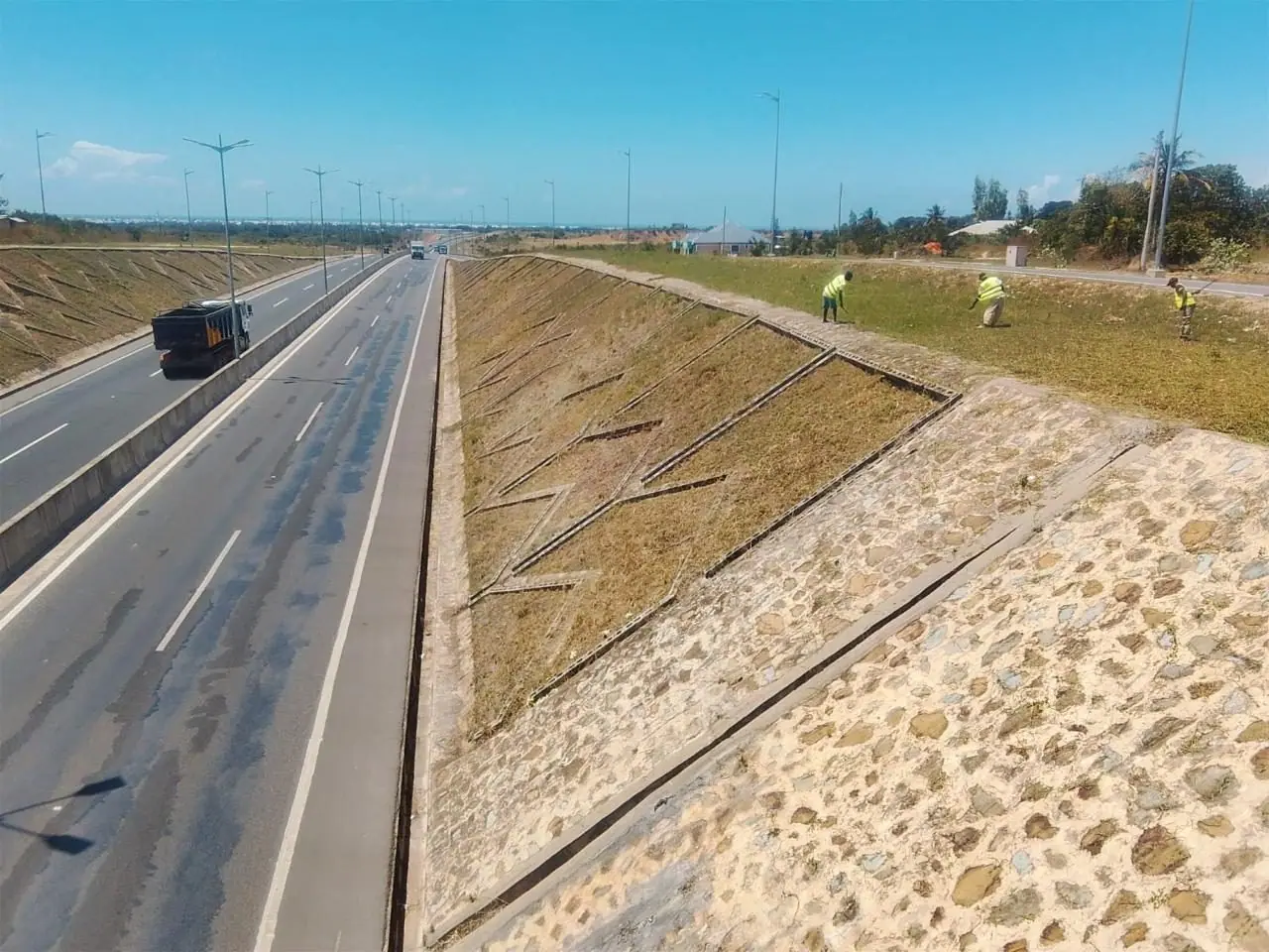 An elevated view of a maintained highway embankment on the Mteza–Kibundani road, with crew tending the verge
